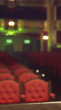 A theater setup shows rows of red seats with soft lighting in the background. The stage is visible, ready for a performance. The atmosphere hints at excitement and anticipation from the audience.
