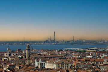 Naklejka premium Historic Venice rooftops and a calm lagoon contrast with distant industrial smokestacks under a hazy sky. A powerful visual for environmental campaigns, urban pollution and climate change.