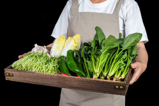 A woman holding a wooden tray filled with fresh vegetables against a black background.