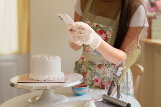 Asian baker writing cake recipe notes while decorating white frosted cake in bright home kitchen workspace