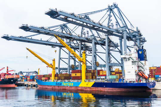 Container ship at a terminal, alongside massive gantry cranes loading and unloading multicolored cargo containers. L&aacute;zaro C&aacute;rdenas, Mexico.