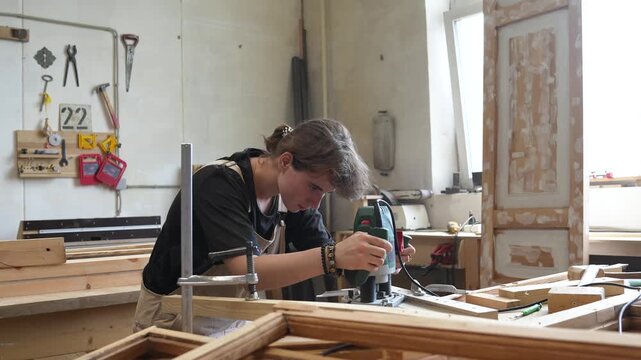 Creative process in woodworking restoration workshop, craftswoman using electric wood router instrument, processing wooden detail, trimming and carving surface of board, handmade craft and renewal.