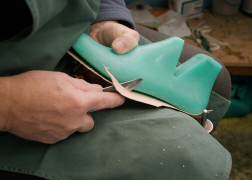 Shoemaker workshop. Handmade footwear. Unrecognizable tailor man hands cutting shaping insole on wooden shoe last using knife at workspace.