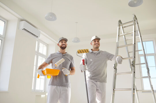 Smiling cheerful team of house painters in uniform standing in empty room near ladder preparing for painting the wall of new apartment. Building contractors doing repair renovation preparing to move.