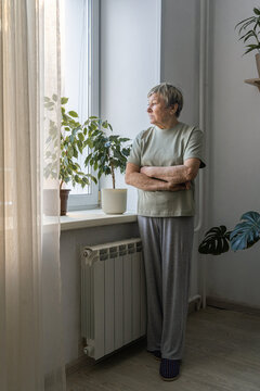 Senior woman standing by window with arms crossed, thoughtful and pensive in quiet home light, reflecting on solitude and memory