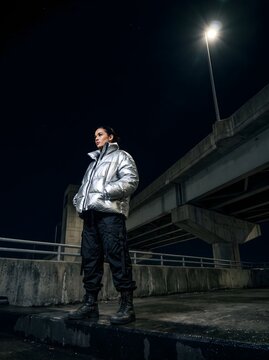 Stylish young woman in a silver puffer jacket standing at night under urban street light