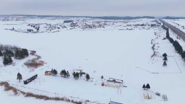 Wide aerial drone footage sweeping over a frozen gravel pit and rural Caledon landscape, Ontario, in deep winter, with snow fields, rustic buildings, winding roads, moody overcast sky.