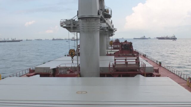 wide high angle view from a bulk carrier ship anchored at kelong anchorage in malaysia, showcasing the main deck, hatch covers, cranes and distant other anchored vessels under a bright cloudy sky