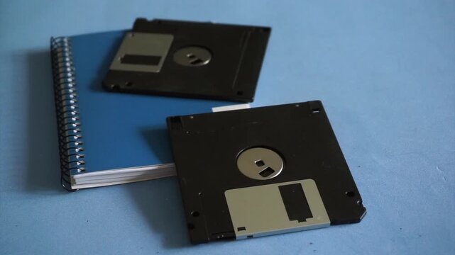 Person's hand placing a black 3.5-inch floppy disk on a blue desk with a spiral notebook