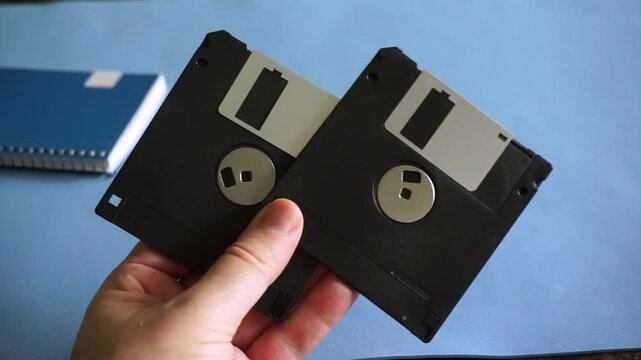 Male hand holding two obsolete 3.5 inch floppy disks over a blue desk with a notebook