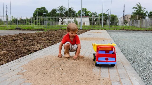 A four-year-old boy with light brown hair, wearing beige boots and shorts, plays with a toy dump truck with a yellow back on a summer day. The boy is playing construction with toy construction equipme