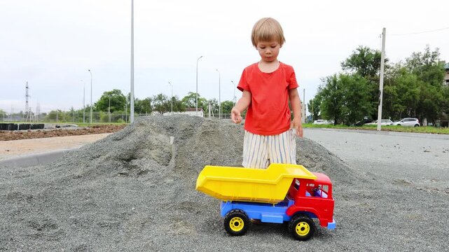 A 4-year-old boy wearing shorts and rubber boots plays with a large toy dump truck on a summer day. He loads the truck bed with gravel using his hands. Active outdoor play of a child