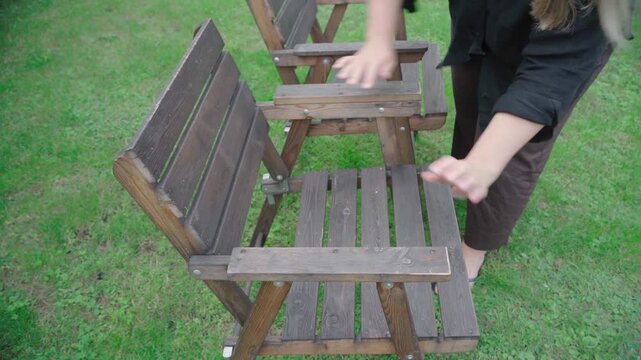 Person in dark clothing rocks a wobbly wooden garden armchair on lawn, testing loose joints and stability for potential repair.