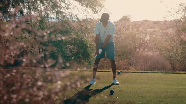 African American golfer hits iron shot on tree lined par 3 tee box at twilight, bright sun flare behind him through pink blooms in slow motion, 4k 800fps golf swing