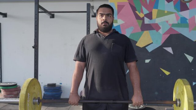 Bearded South Asian male athlete performs a barbell deadlift during functional fitness training while the camera moves smoothly side to side from a front view. Collection: WORKOUTSERIES2026