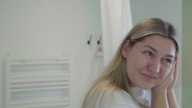 Adult holds both hands to her ear in a bathroom, wincing slightly and checking her reflection as if feeling ear pain or pressure.