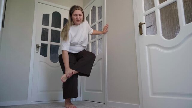 Barefoot woman in a hallway stops, balances on one leg and clutches her aching foot after walking on the carpet.