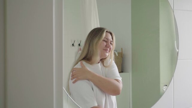 In a bathroom mirror reflection, a tired-looking home dweller grips her upper arm and shoulder, wincing from muscle ache.