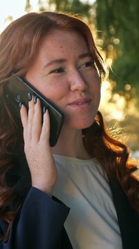 Beautiful red haired woman with freckles talking on her smartphone in a park. Warm sunset light illuminates her face and curly hair as she smiles, enjoying a pleasant conversation outdoors