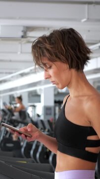 Athletic woman with short hair browsing her smartphone while walking on a treadmill in a fitness club, focused on her device during a cardio workout session to stay connected and entertained