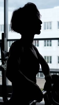 Unrecognizable woman bodybuilder doing an intense workout with a barbell in front of a window, showcasing her strength and muscular physique while performing weightlifting exercises in a gym