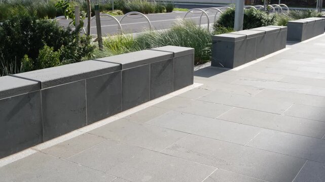Stone block benches with installed metal skate deterrents along a paved pedestrian concourse in a modern public plaza. In the background, hoops for bicycle parking line the street in Australia