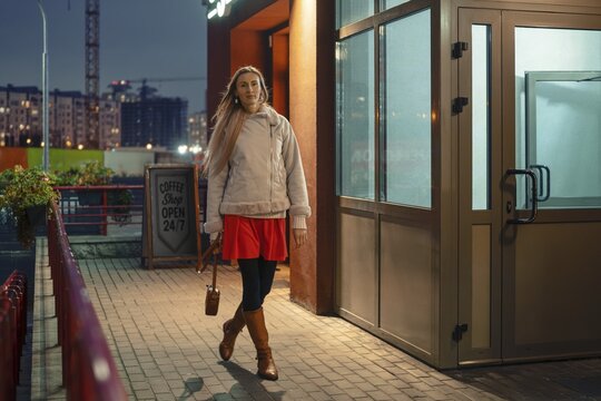 A woman strolls outside a coffee shop in the evening. She is dressed in a bright red dress, brown boots, and a cozy jacket