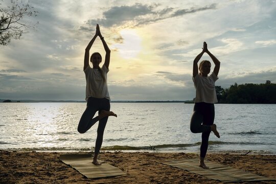 Silhouettes of two women performing yoga poses on mats along a sandy lakeshore as the sun sets, casting a warm glow over the water and creating a peaceful atmosphere