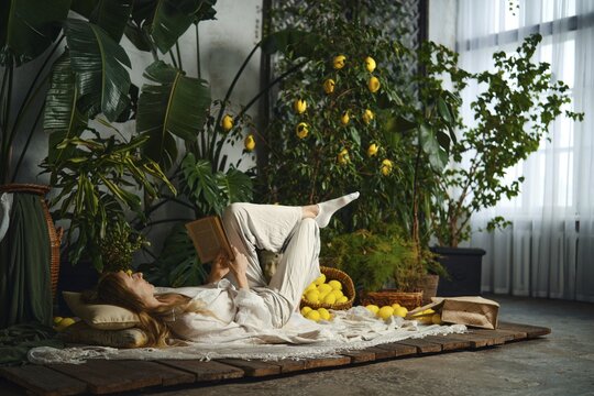 A person lies on the floor and reads a book with her feet in the air. The room is filled with green plants and baskets of lemons, creating calm and relaxed atmosphere. Natural light comes through windows