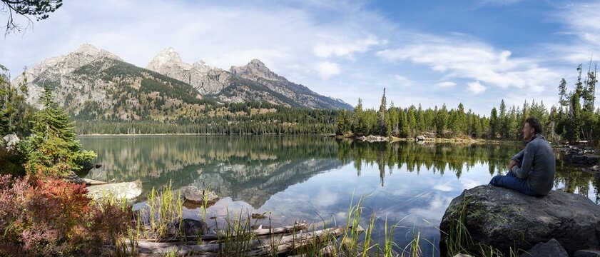 Young man sitting on a stone by a lake, reflection in Taggart Lake, view of Teton Range mountain, summit Grand Teton and Teewinot Mountain, Grand Teton National Park, Wyoming, USA