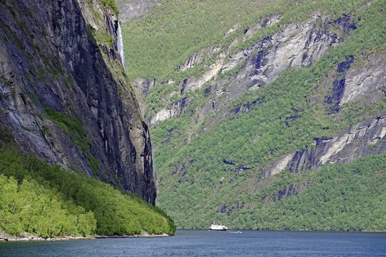 A boat sails through a fjord surrounded by steep green cliffs under a blue sky with scattered clouds