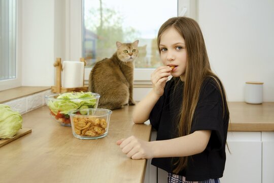 Cat is watching how little girl bites and crunches a cracker while standing in the kitchen