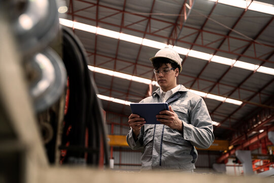 Asian Male Engineer in Gray Uniform Using Tablet While Standing at Industrial Production Line. Digital Manufacturing Operations and Smart Factory Professional.