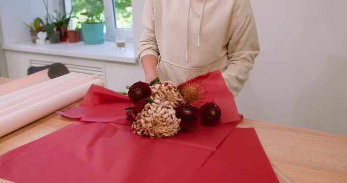 Bouquet presentation. Flowers packaging. Unrecognizable professional woman florist using red paper for dahlia on table in workshop.