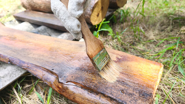 Close-up of paintbrush coating weathered wood with protective stain. Wood restoration, outdoor carpentry, DIY renovation and maintenance project.