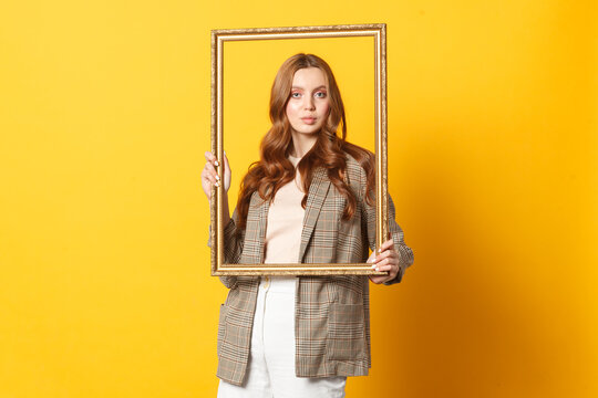 Young woman holding an empty picture frame against a yellow background. Creativity, identity and self-expression concept.