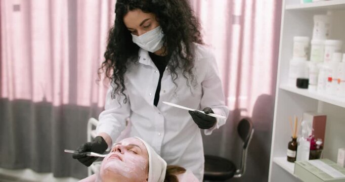 Cosmetologist working. Facial service. Woman in gloves with brush applying white cream for professional mask using spatula in spa clinic.