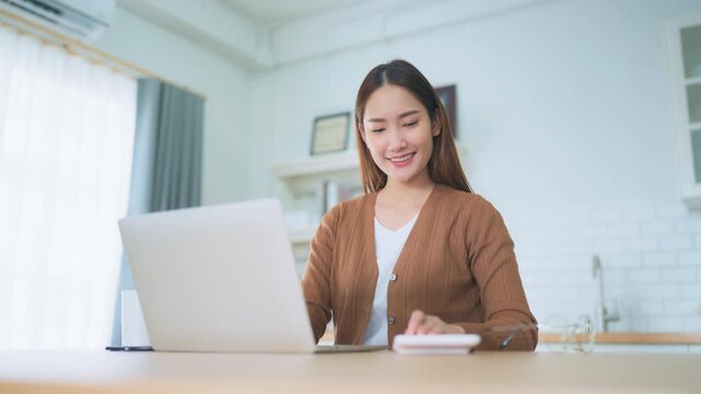 Beautiful young Asian woman working with laptop computer in home kitchen. Work at home
