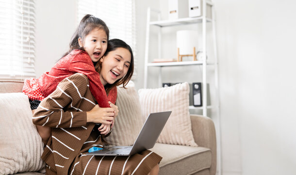 Cheerful Asian mother carrying daughter piggyback while working on laptop at home