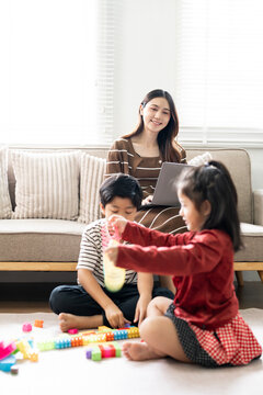 Asian children playing together on floor with mother working on laptop in living room
