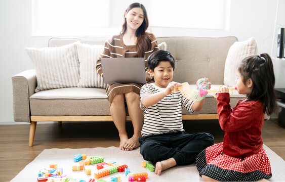 Asian brother and sister playing with toys on floor while mother working on laptop on sofa