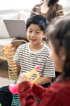 Asian boy playing with slinky toy cheerfully with his sister and working mother in background