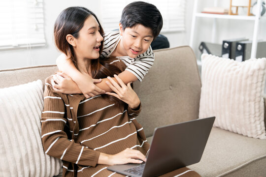 Happy Asian mother working on laptop while her loving son embraces her from behind at home