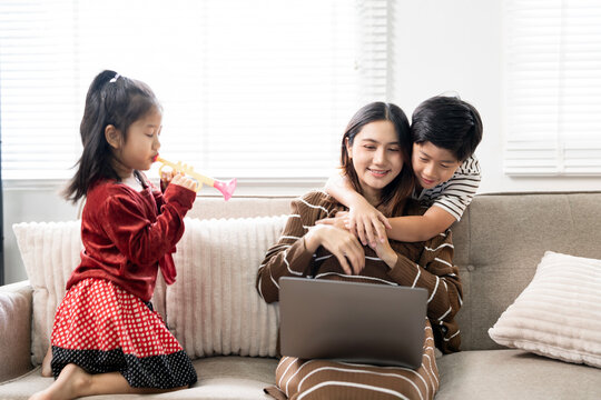 Happy Asian mother sitting on sofa with laptop while son embraces her and daughter plays toy trumpet