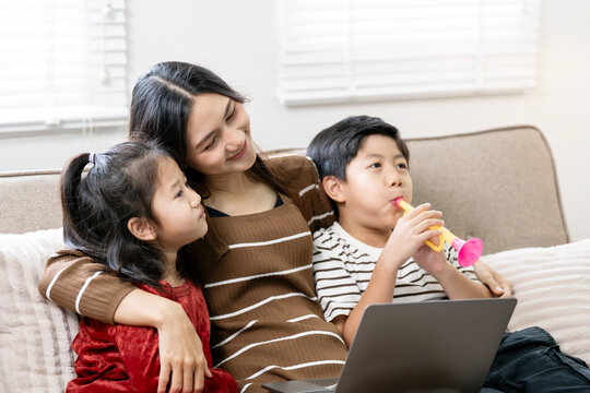 Happy Asian mother hugging daughter and sitting with son playing toy trumpet on sofa at home