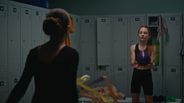 Female Gymnast Practicing Complex Coordination in Dim Locker Room. Girl Handles Two Sports Apparatuses Simultaneously, Creating Colorful Spirals With Ribbon While Focusing Eyes to Catch Purple Ball.