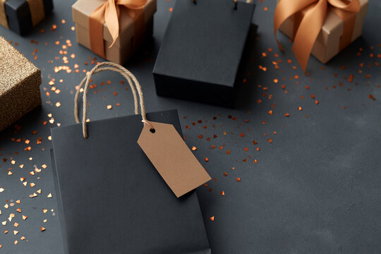 Black gift bag sits on dark textured surface. Brown tag hangs from twine handle, minimalist and elegant. Golden confetti scatters around, adding festive sparkle