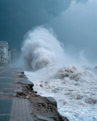 Fototapeta premium Stormy sea crashes against broken coastal walkway. Foam explodes under dark, threatening skies. Broken tiles and rubble mark erosion's fury. Distant buildings stand silent, battered by wind