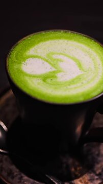 Matcha latte green tea with milk foam heart art in black cup on dark table background camera zoom in.