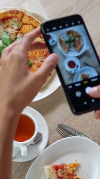 Woman's hands using a mobile phone to take a picture of a fresh italian pizza on a wooden table in a cafe, capturing food for social media or a blog before starting her meal